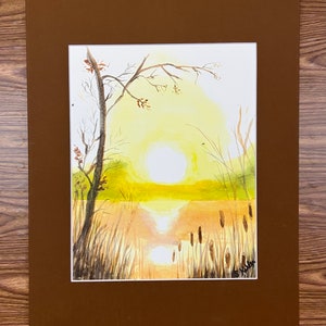 May include: Watercolor painting of a sunset over a lake, with tall grasses and trees in silhouette. The sun is a bright yellow orb, reflecting on the water. The artwork is framed in a brown mat.