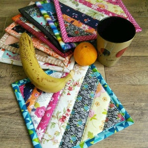 Peut inclure: Un ensemble de maniques en tissu coloré avec un motif de patchwork diagonal. Les maniques sont faites de différents tissus avec des motifs floraux et géométriques. Une banane, une orange et une tasse à café sont sur la table à côté des maniques.