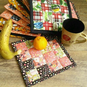 May include: A set of colorful patchwork fabric pot holders with a black trim. The pot holders are made of different fabrics with floral and geometric patterns. A mug and an orange are on top of one of the pot holders.