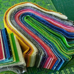 May include: A large stack of folded fabric strips in a rainbow of colours including yellow, orange, red, pink, purple, blue, and green. The fabric is arranged in a curved, layered formation, with a smaller, separate stack of fabric strips nearby.