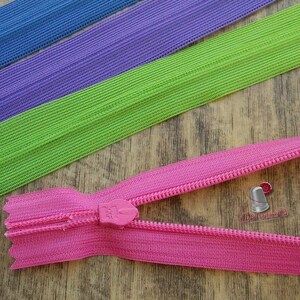 Pu&ograve; includere: Una cerniera lampo rosa con un cursore in plastica. La cerniera &egrave; aperta e i denti sono visibili. La cerniera &egrave; appoggiata su una superficie di legno. Sullo sfondo ci sono tre strisce di tessuto elastico: blu, viola e verde.