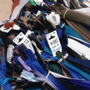 Pu&ograve; includere: Un vasto assortimento di cerniere lampo di diversi colori, tra cui bianco, nero, blu, marrone e blu-verde. Le cerniere lampo sono raggruppate e legate con uno spago. Un'etichetta di prezzo per le cerniere lampo "Invisible 4408-08" &egrave; visibile.