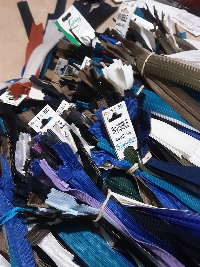 Pu&ograve; includere: Un vasto assortimento di cerniere lampo di diversi colori, tra cui bianco, nero, blu, marrone e blu-verde. Le cerniere lampo sono raggruppate e legate con uno spago. Un'etichetta di prezzo per le cerniere lampo "Invisible 4408-08" &egrave; visibile.