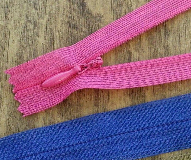 Pu&ograve; includere: Due nastri di cerniera, uno rosa e uno blu, sono mostrati su una superficie di legno. Il nastro di cerniera rosa ha un cursore rosa.