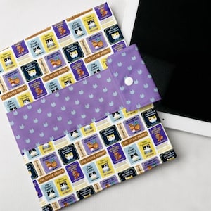 Pode incluir: Uma capa de livro de tecido roxo e branco com um padrão repetido de gatos e títulos de livros. A capa tem uma aba com fecho de botão. Os títulos dos livros incluem "The Great Gatsby", "Oliver's Adventures in Wonderland" e "The Litter Prince".