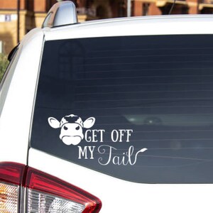K&ouml;nnte beinhalten: Wei&szlig;es Auto mit einem schwarzen Kuh-Aufkleber auf der Heckscheibe. Der Aufkleber lautet "Get Off My Tail".