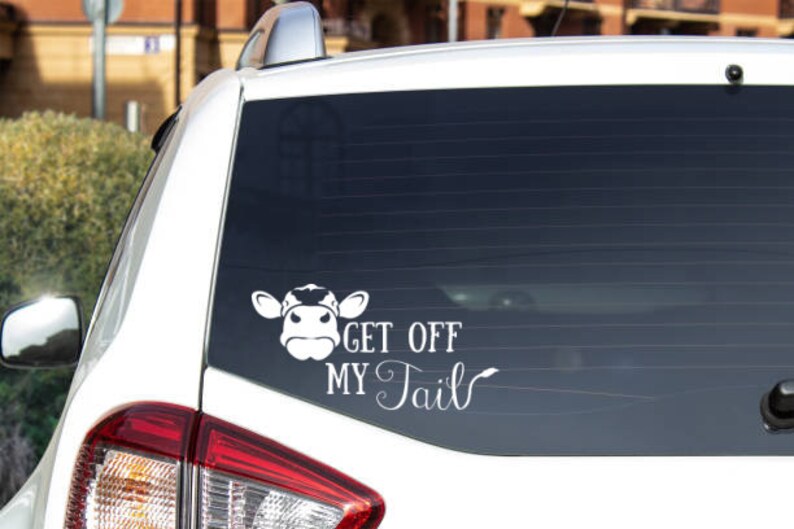 K&ouml;nnte beinhalten: Wei&szlig;es Auto mit einem schwarzen Kuh-Aufkleber auf der Heckscheibe. Der Aufkleber lautet "Get Off My Tail".