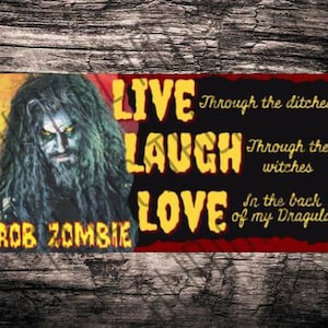 May include: A black and red rectangular sign with the words "LIVE LAUGH LOVE" in yellow text. The text is surrounded by a black border. The sign also features a portrait of Rob Zombie in the upper left corner and the text "Through the ditches", "Through the witches", and "In the back of my Dragula" in yellow text.
