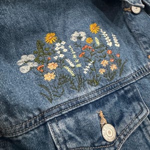 May include: Blue denim jacket with embroidered floral design in yellow, white, orange, and green. The jacket has two front pockets and silver buttons. The embroidery is on the upper left chest area.