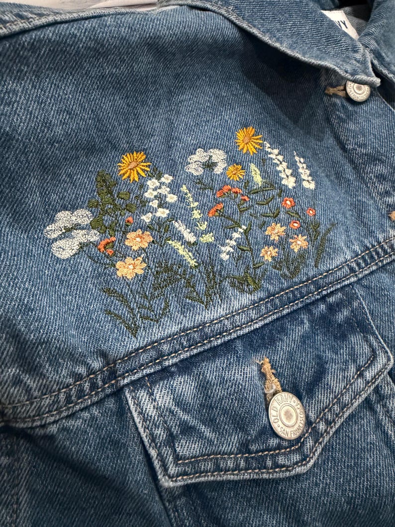 May include: Blue denim jacket with embroidered floral design in yellow, white, orange, and green. The jacket has two front pockets and silver buttons. The embroidery is on the upper left chest area.
