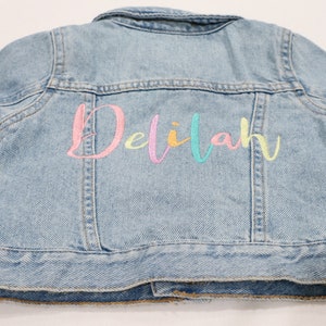 K&ouml;nnte beinhalten: Eine hellblaue Jeansjacke mit dem Namen "Delilah" in Regenbogenfarben auf der R&uuml;ckseite gestickt.