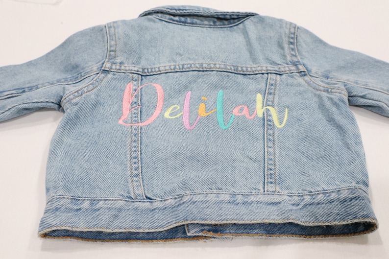 K&ouml;nnte beinhalten: Eine hellblaue Jeansjacke mit dem Namen "Delilah" in Regenbogenfarben auf der R&uuml;ckseite gestickt.
