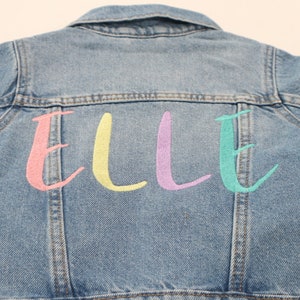 K&ouml;nnte beinhalten: Eine hellblaue Jeansjacke mit dem Namen "ELLE" in Pink, Gelb, Lila und T&uuml;rkis auf der R&uuml;ckseite gestickt.