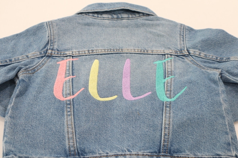 K&ouml;nnte beinhalten: Eine hellblaue Jeansjacke mit dem Namen "ELLE" in Pink, Gelb, Lila und T&uuml;rkis auf der R&uuml;ckseite gestickt.