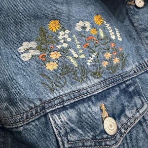May include: Close-up of a blue denim jacket with embroidered floral design. The embroidery features yellow, white, and orange flowers with green stems and leaves. The jacket has a button closure and a chest pocket.