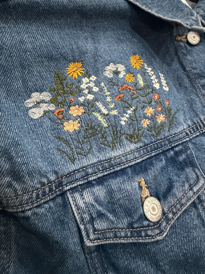 May include: Close-up of a blue denim jacket with embroidered floral design. The embroidery features yellow, white, and orange flowers with green stems and leaves. The jacket has a button closure and a chest pocket.