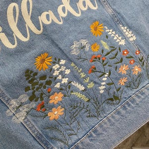 May include: Light blue denim jacket with embroidered floral design in yellow, orange, white, and green. The jacket features the name "Mrs. Illada" in white script across the back, making it a personalized item.