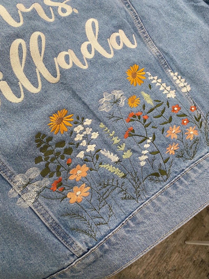 May include: Light blue denim jacket with embroidered floral design in yellow, orange, white, and green. The jacket features the name "Mrs. Illada" in white script across the back, making it a personalized item.