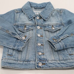 K&ouml;nnte beinhalten: Eine hellblaue Jeansjacke mit Knopfleiste und zwei Fronttaschen. Die Jacke hat den Schriftzug "Old Navy" auf der Innenseite des Kragens.
