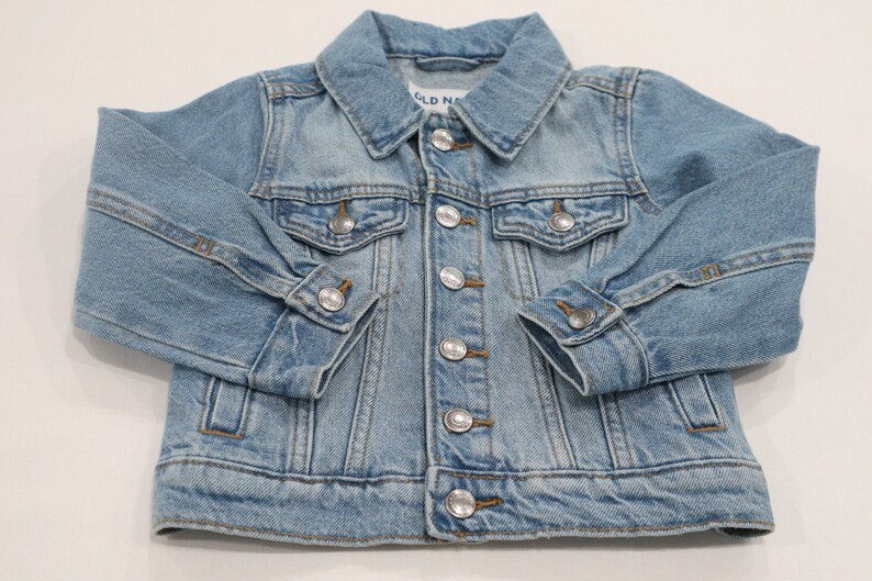 K&ouml;nnte beinhalten: Eine hellblaue Jeansjacke mit Knopfleiste und zwei Fronttaschen. Die Jacke hat den Schriftzug "Old Navy" auf der Innenseite des Kragens.