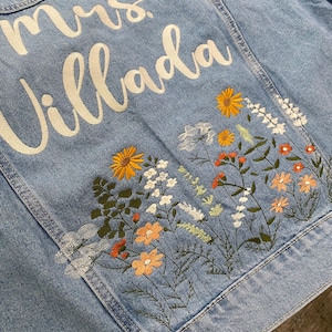 May include: Light blue denim jacket with embroidered floral design in orange, yellow, and white. The jacket features the text "mrs. Villada" in white script across the back. A stylish and personalized piece.