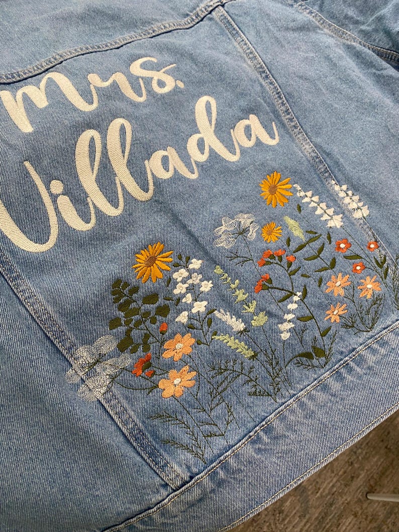 May include: Light blue denim jacket with embroidered floral design in orange, yellow, and white. The jacket features the text "mrs. Villada" in white script across the back. A stylish and personalized piece.