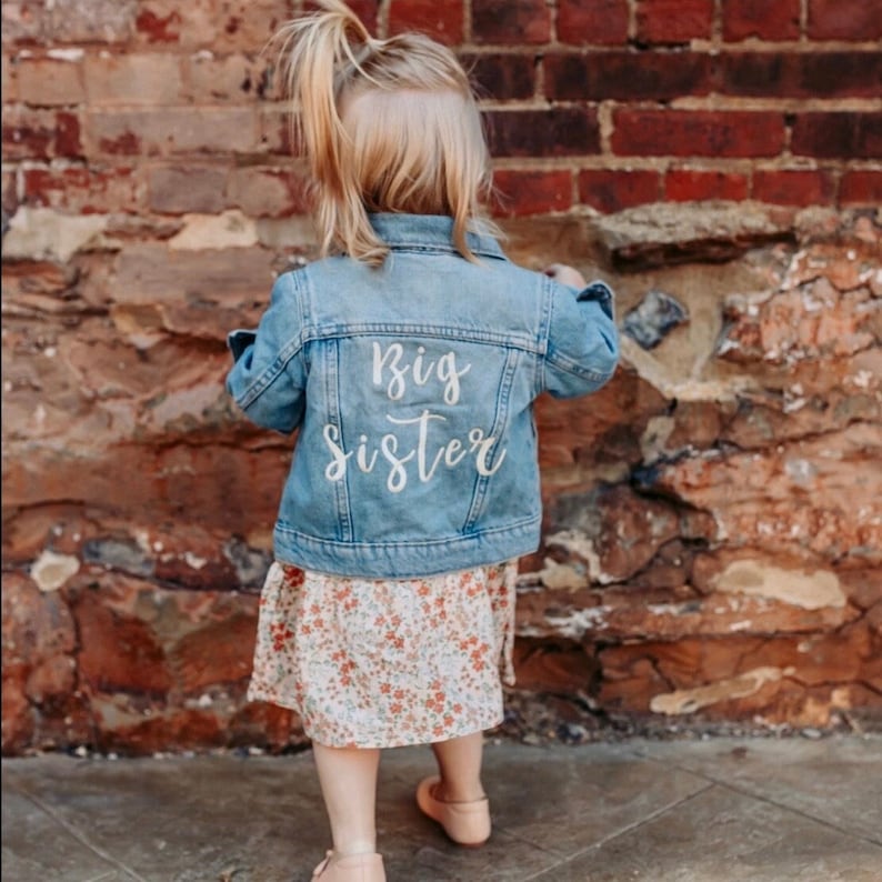 May include: A young child wearing a light blue denim jacket with the words "Big Sister" written in white on the back. The child is wearing a floral dress and is standing in front of a brick wall.