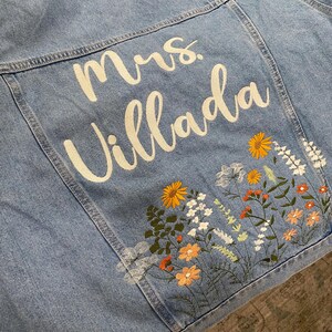 May include: Light blue denim jacket with embroidered floral design and the name "Mrs. Villada" in white script. The floral embroidery includes orange, yellow, and white flowers with green leaves. The jacket is a classic style.