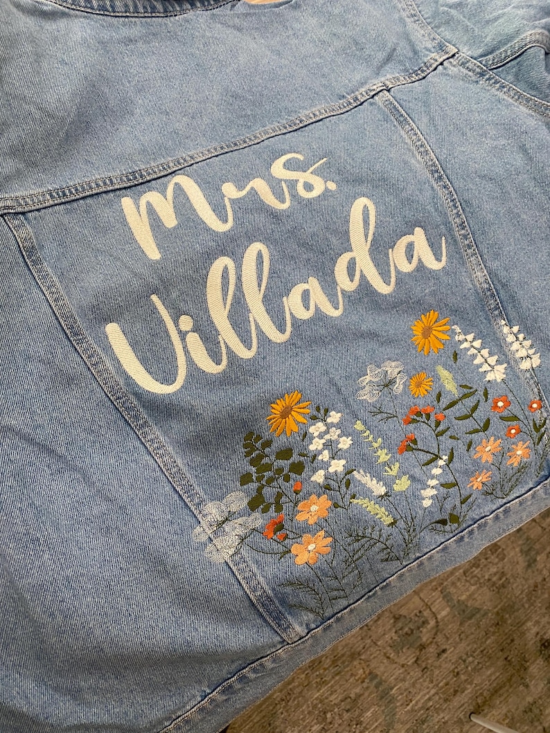 May include: Light blue denim jacket with embroidered floral design and the name "Mrs. Villada" in white script. The floral embroidery includes orange, yellow, and white flowers with green leaves. The jacket is a classic style.