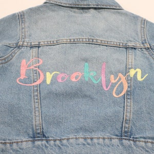 K&ouml;nnte beinhalten: Eine hellblaue Jeansjacke mit dem Namen "Brooklyn", der in Regenbogenfarben auf der R&uuml;ckseite gestickt ist.