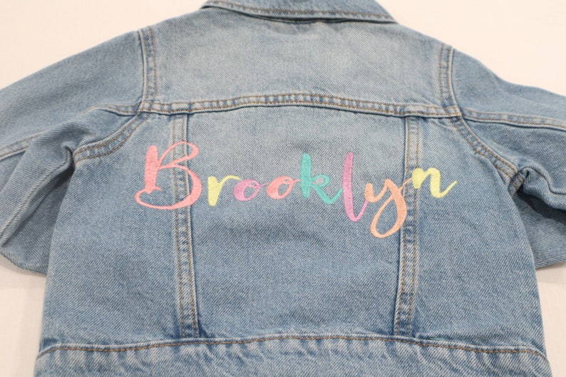 K&ouml;nnte beinhalten: Eine hellblaue Jeansjacke mit dem Namen "Brooklyn", der in Regenbogenfarben auf der R&uuml;ckseite gestickt ist.