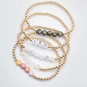 May include: Four gold bead bracelets with different names spelled out in colorful beads. The names are James, Leo, Claire, and Navy.