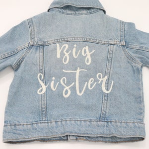 May include: A light blue denim jacket with the words "Big Sister" embroidered on the back in white.