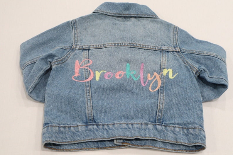K&ouml;nnte beinhalten: Eine hellblaue Jeansjacke mit dem Namen "Brooklyn", der in Regenbogenfarben auf der R&uuml;ckseite gestickt ist.