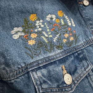 May include: Close-up of a denim jacket with embroidered floral design. The jacket is a medium wash blue with a detailed floral pattern in yellow, white, orange, and green thread. Features include a button closure and two front pockets.