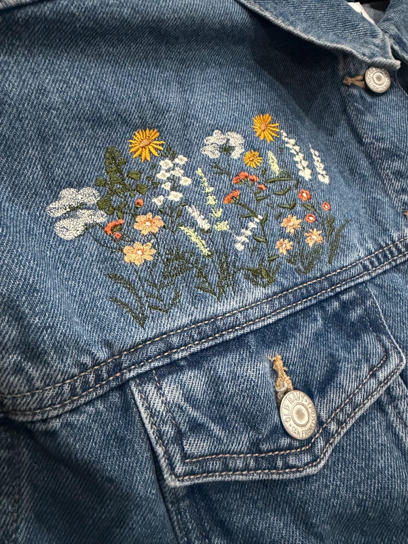May include: Close-up of a denim jacket with embroidered floral design. The jacket is a medium wash blue with a detailed floral pattern in yellow, white, orange, and green thread. Features include a button closure and two front pockets.