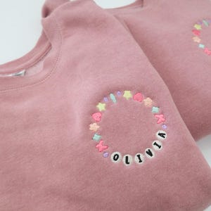 Könnte beinhalten: Zwei rosafarbene Sweatshirts mit gestickten Kreisen aus bunten Sternen und den Namen "Olivia" und "Luca" in Schwarz.
