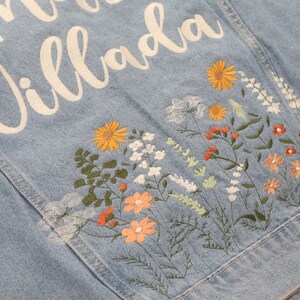 May include: Light blue denim jacket with embroidered floral design. The back of the jacket features the name "Villada" in white script. The floral embroidery includes orange, yellow, and white flowers with green leaves.