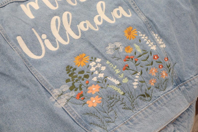 May include: Light blue denim jacket with embroidered floral design. The back of the jacket features the name "Villada" in white script. The floral embroidery includes orange, yellow, and white flowers with green leaves.