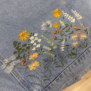 May include: Light blue denim jacket with embroidered floral design. The embroidery features yellow, orange, and white flowers with green leaves. The jacket has a classic denim design with pockets and seams.