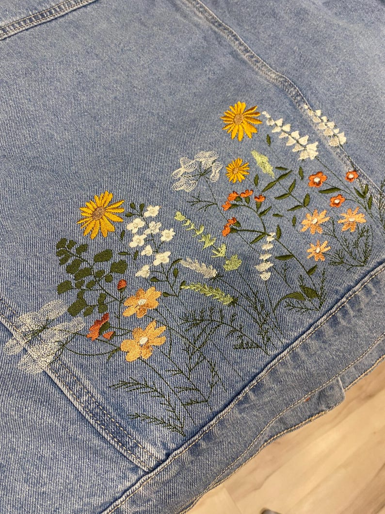 May include: Light blue denim jacket with embroidered floral design. The embroidery features yellow, orange, and white flowers with green leaves. The jacket has a classic denim design with pockets and seams.