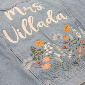 May include: Light blue denim jacket with white script text that reads "Mrs. Villada". The jacket features embroidered flowers in shades of orange, yellow, white, and green. A stylish and personalized denim jacket.