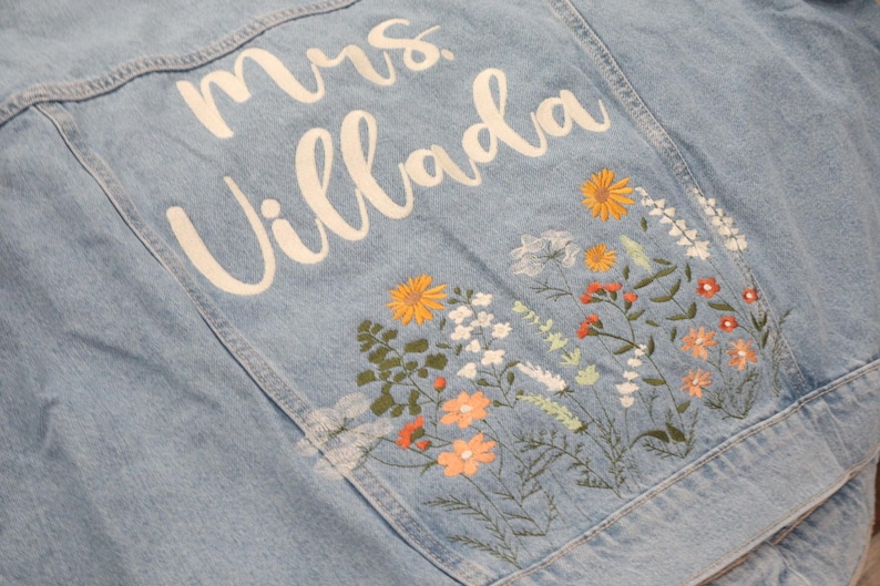 May include: Light blue denim jacket with white script text that reads "Mrs. Villada". The jacket features embroidered flowers in shades of orange, yellow, white, and green. A stylish and personalized denim jacket.