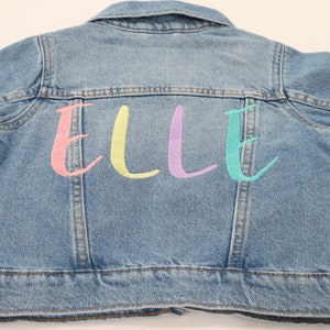 K&ouml;nnte beinhalten: Eine hellblaue Jeansjacke mit dem Namen "ELLE" in rosa, gelb, lila und t&uuml;rkis auf der R&uuml;ckseite gestickt.