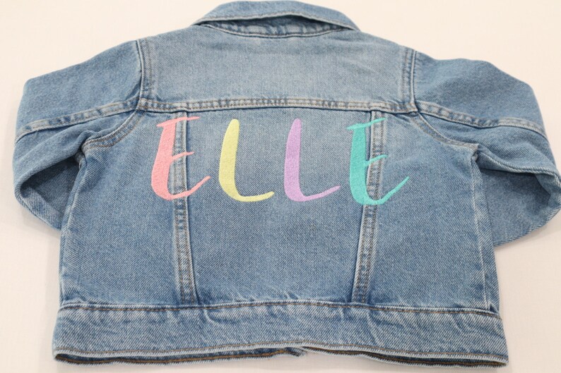K&ouml;nnte beinhalten: Eine hellblaue Jeansjacke mit dem Namen "ELLE" in rosa, gelb, lila und t&uuml;rkis auf der R&uuml;ckseite gestickt.