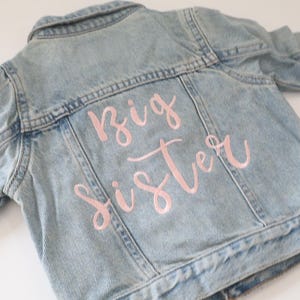 May include: A light blue denim jacket with the words "Big Sister" embroidered in pink on the back.
