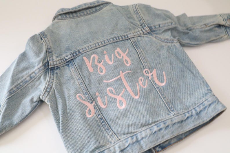 May include: A light blue denim jacket with the words "Big Sister" embroidered in pink on the back.