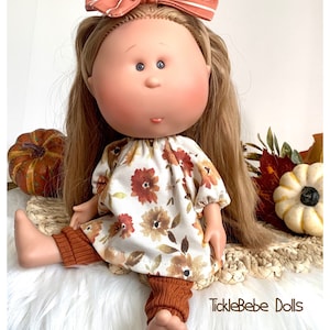 May include: A doll with long blonde hair, blue eyes, and rosy cheeks, wearing a floral print dress and rust-colored leggings. A rust-colored bow adorns the doll's head. The doll is sitting on a woven mat with pumpkins and leaves in the background. The text "TickleBebe Dolls" is visible.