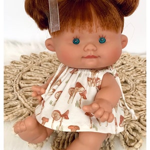 May include: A small doll with red hair in pigtails wearing a white dress with a mushroom print. The doll has blue eyes and is sitting on a woven rug. The text "TickleBebe Dolls" is visible in the bottom right corner of the image.