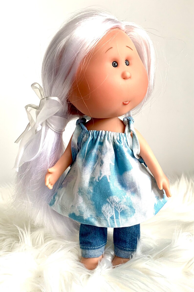 May include: A doll with long white hair, wearing a blue and white patterned dress and denim pants. The doll has large eyes and a sweet expression.